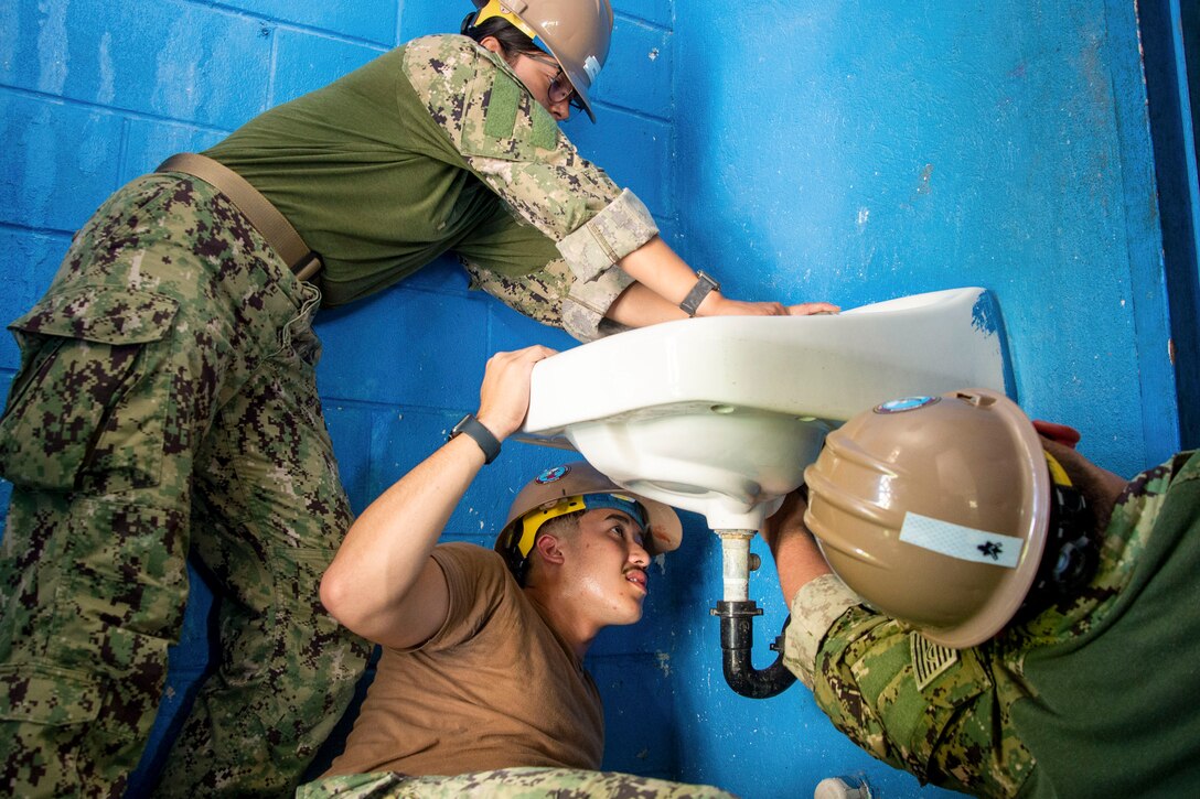 A sailor holds a sink in place as two sailors laying on the ground work on the bottom on it.