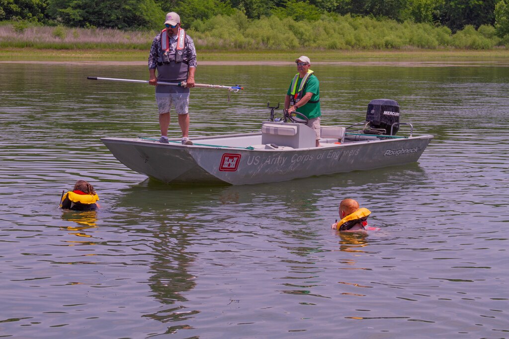 Man stands at edge of boat while other man pilots boat with two swimmers in the water wearing inflatable life jackets.