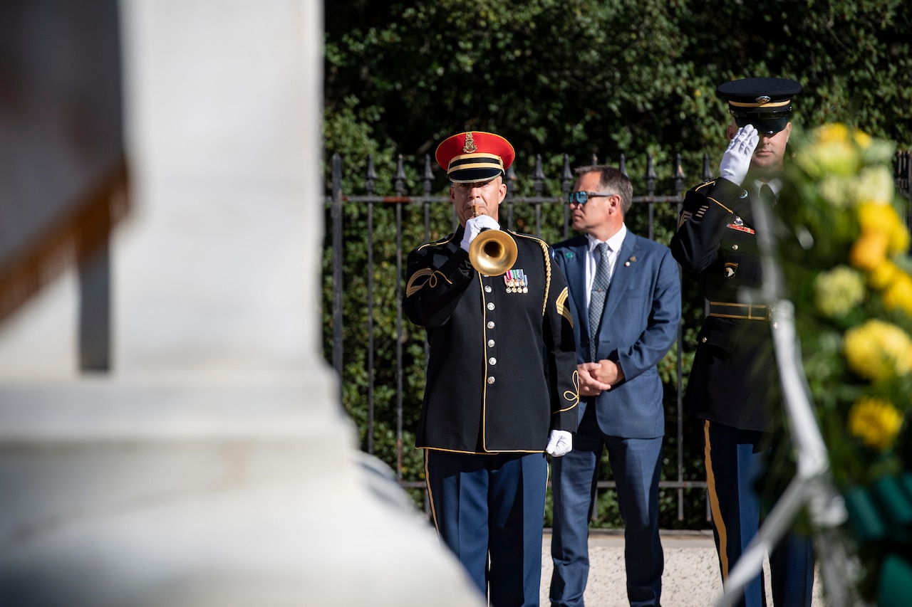 A soldier in in dress uniform with a red hat plays a bugle next to a service member in dress uniform with a blue hat salutes a wreath in the foreground. A person in a suit is in the background.