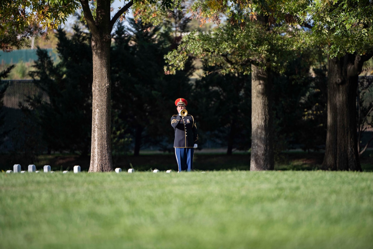 A solider in dress uniform plays the bugle in the background in front of trees by graves of fallen service members.
