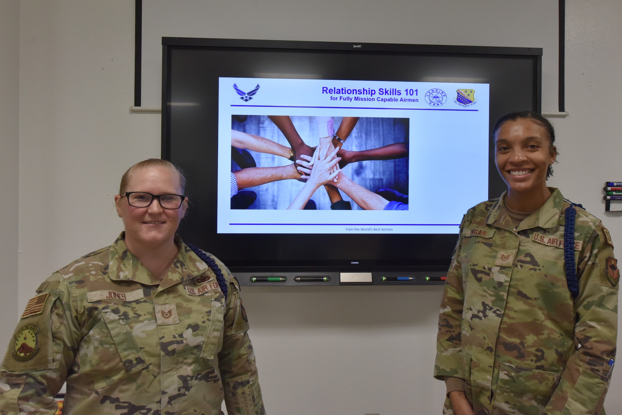 Two Airmen stand in front of a television screen.
