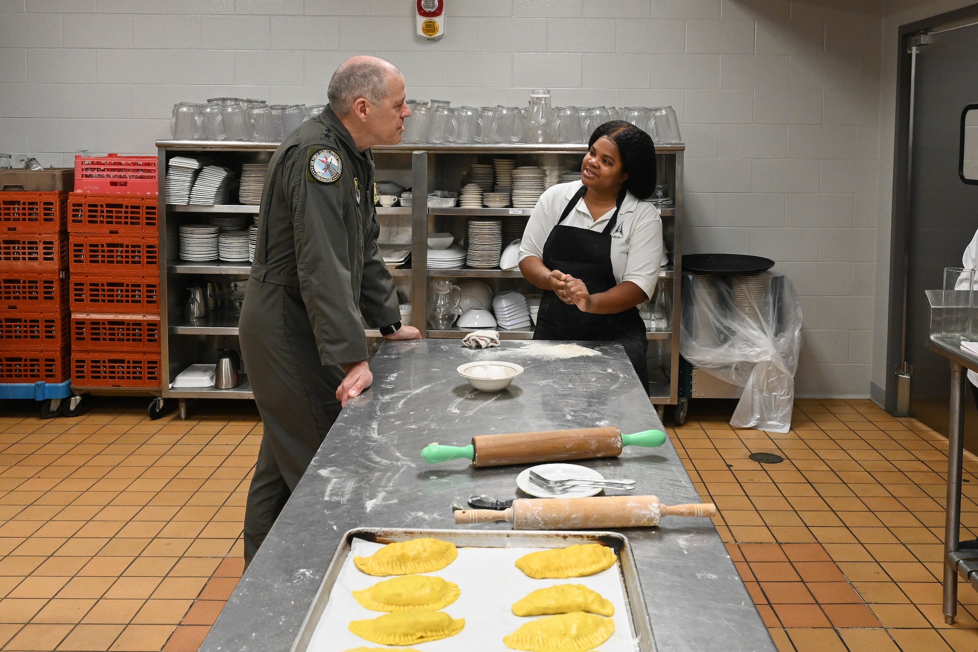 Gen. Tom Bussiere and TSgt. Shameka Risch talk in a kitchen by a table with rolling pins and flour.
