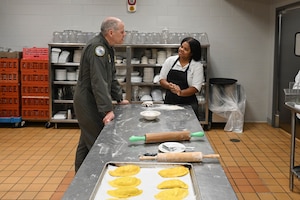 Gen. Tom Bussiere and TSgt. Shameka Risch talk in a kitchen by a table with rolling pins and flour.