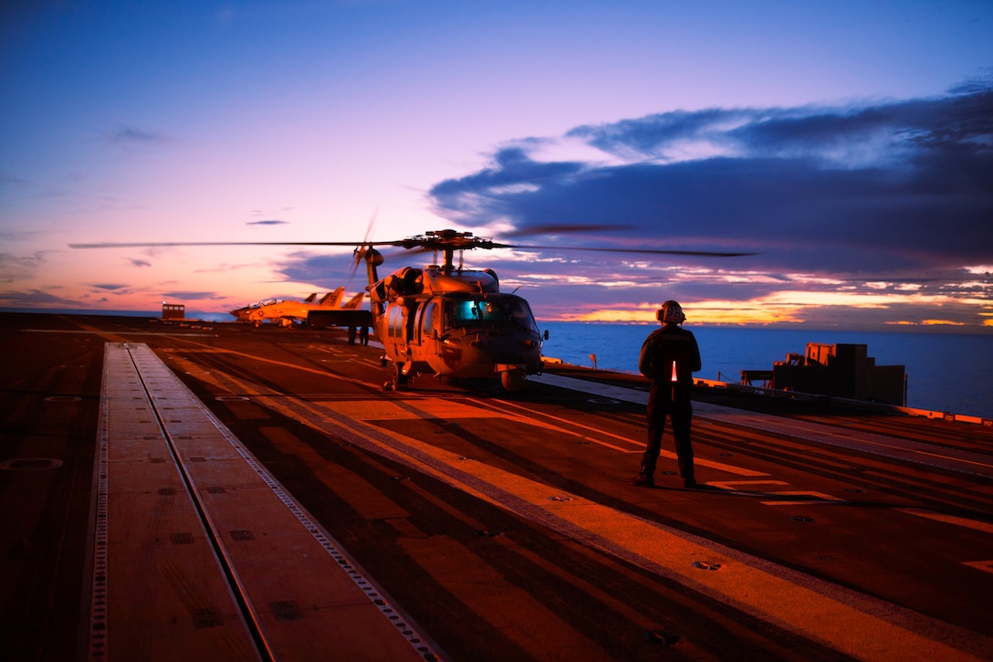 A sailor faces a helicopter on the flight deck of a Navy ship at dawn.