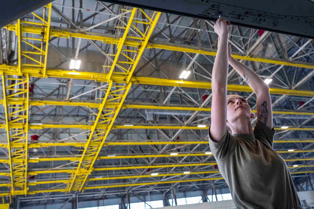 An airman works on an aircraft underneath an industrial ceiling with yellow framework.