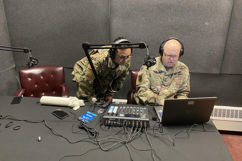 Two soldiers wearing headphones work at a table with recording equipment.