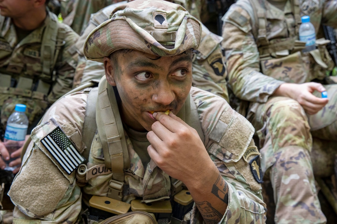 A soldier wearing camouflage kneels while eating in front of fellow soldiers.