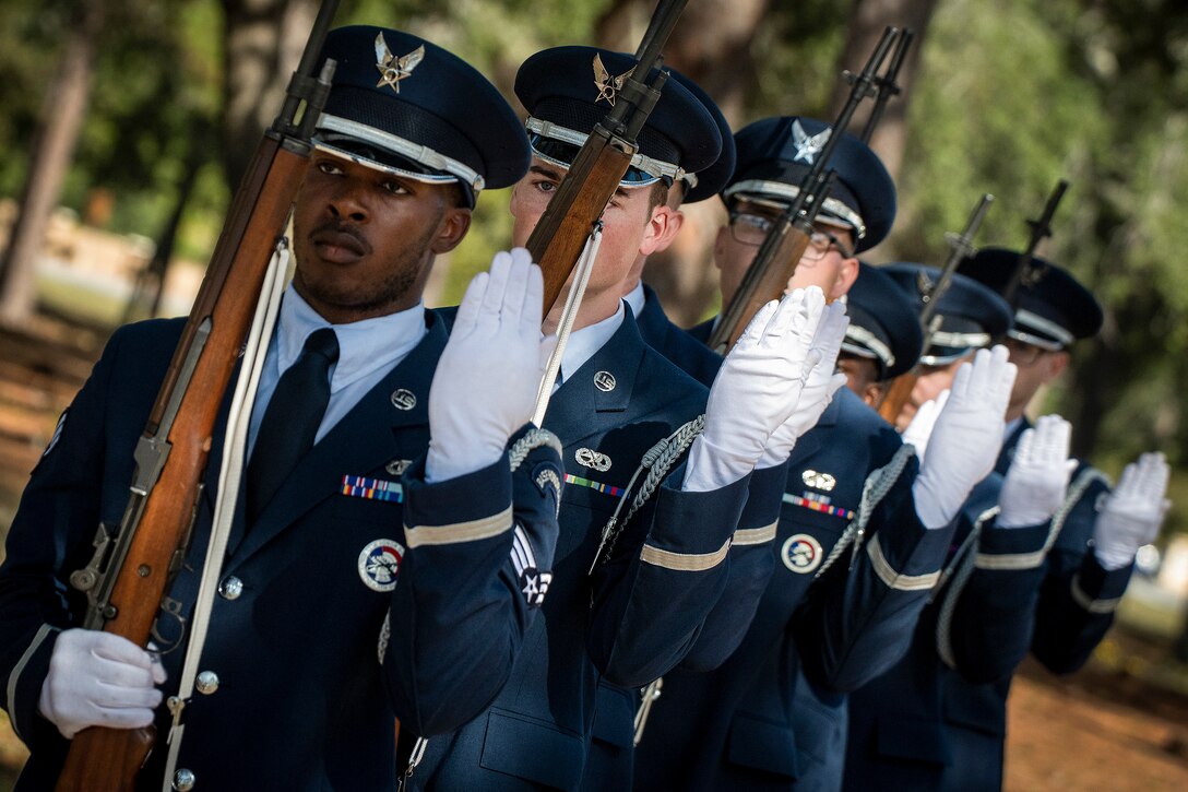 A slanted angle of airmen in ceremonial uniforms standing in formation while holding rifles with blurred trees in the background.