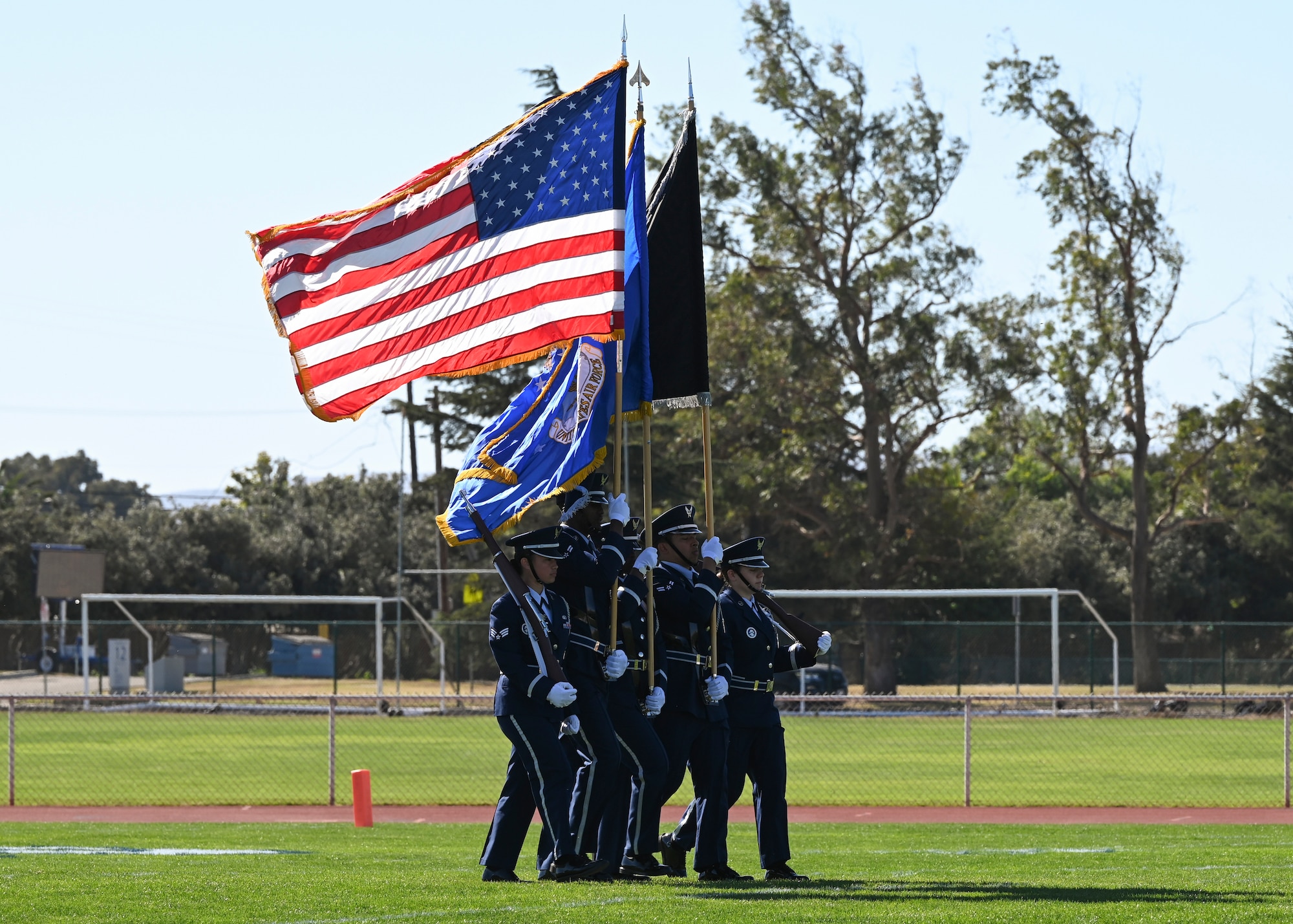 Five service members in blues carry flags and rifles onto a football field.