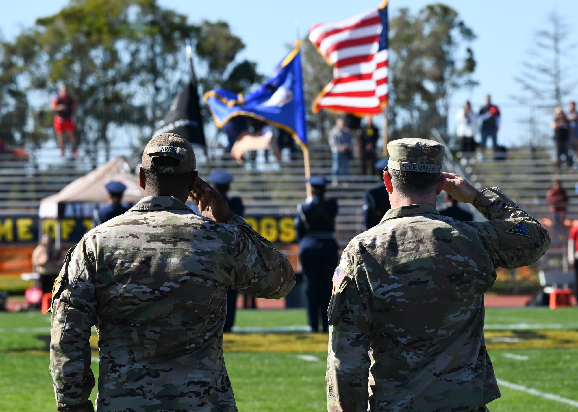 Two service members are saluting flags carried by service members in uniform on a football field.