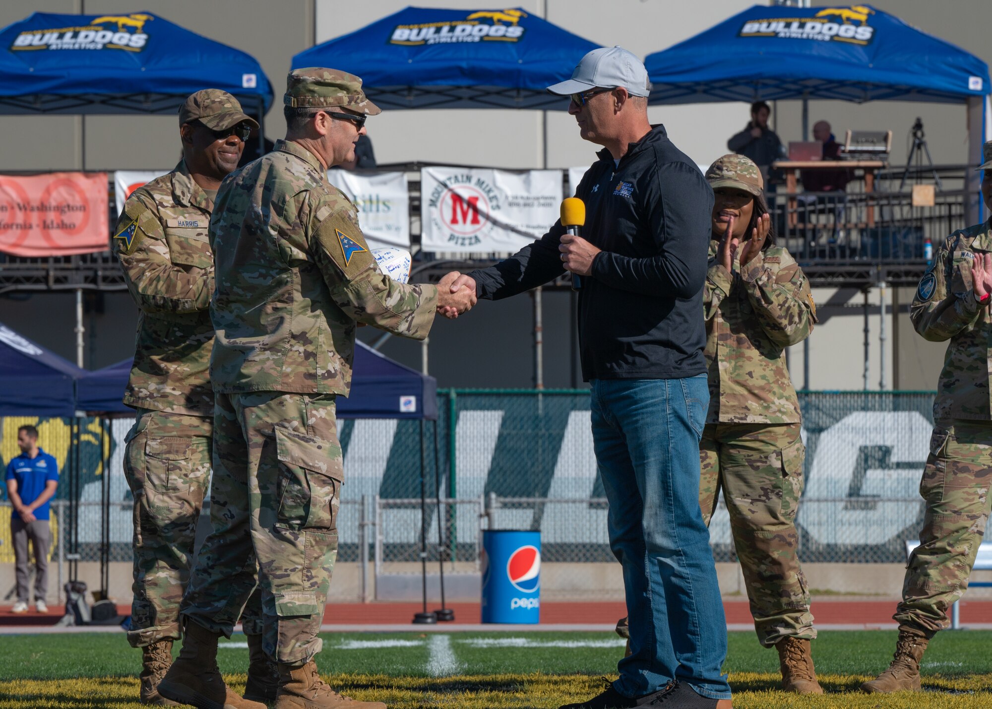 Two people are shaking hands with three service members in the back on a football field.