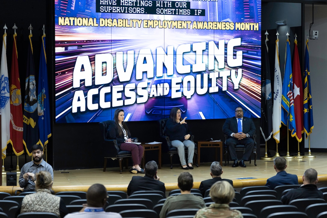 Two light skinned women, one with glasses, sit on stage with a dark skinned man with glasses. The middle woman wearing glasses performs sign language. A audience in an auditorium listens.