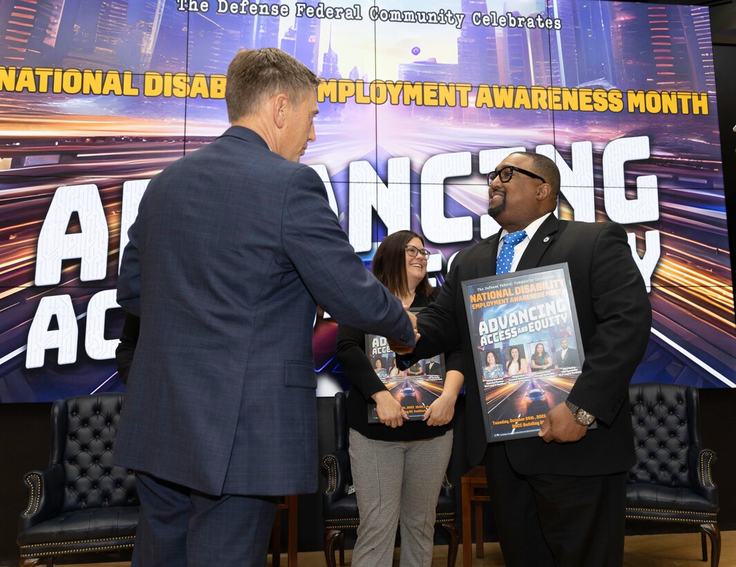 A light skinned man with graying hair shakes the hand of a dark skinned man with glasses on a stage. A light skinned woman with glasses stands in the background. All are in business attire.