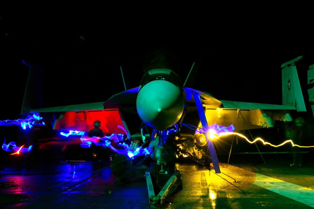 Night view of a military aircraft on the flight deck of a Navy ship. The flight deck is illuminated by red and yellow lights.