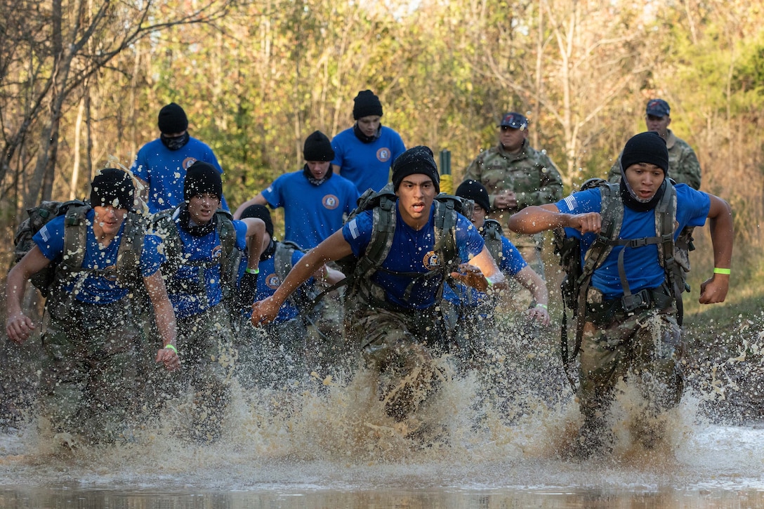 Army JROTC cadets run through water in a wooded area.