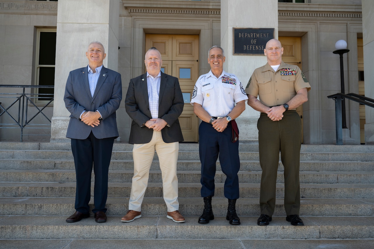 Four men stand on the steps of a building. The sign on the building indicates that they are at the Department of Defense.