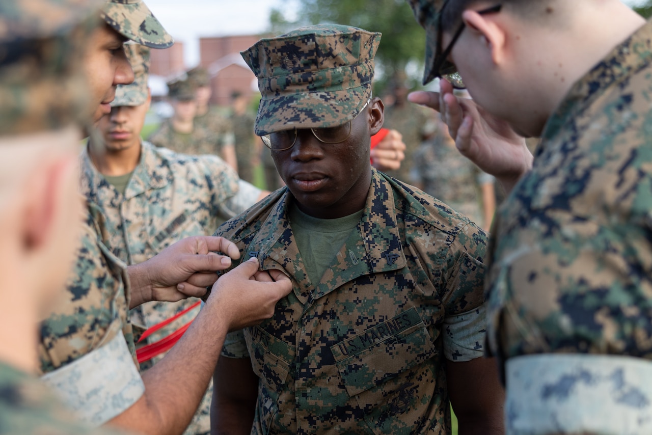 A Marine affixes a pin to another Marine's uniform as others stand by outdoors.