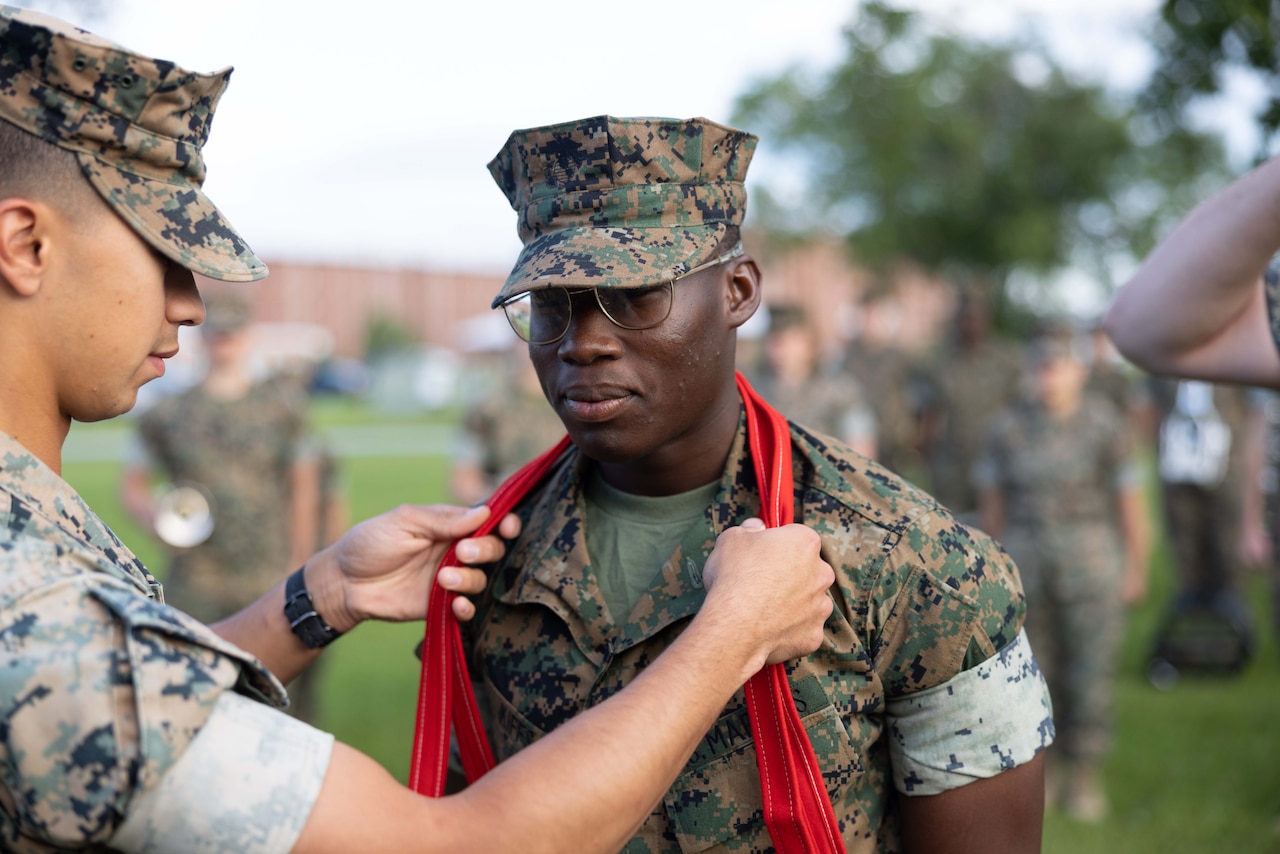 A Marine places a red scarf around the neck of another Marine as others stand in formation outdoors.