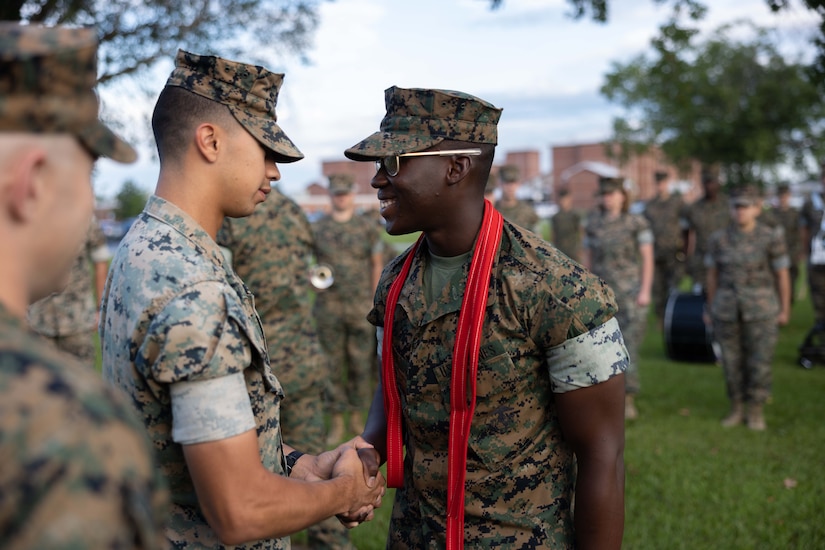A smiling Marine wearing a red scarf with his uniform shakes hands with another Marine as others stand by outdoors.