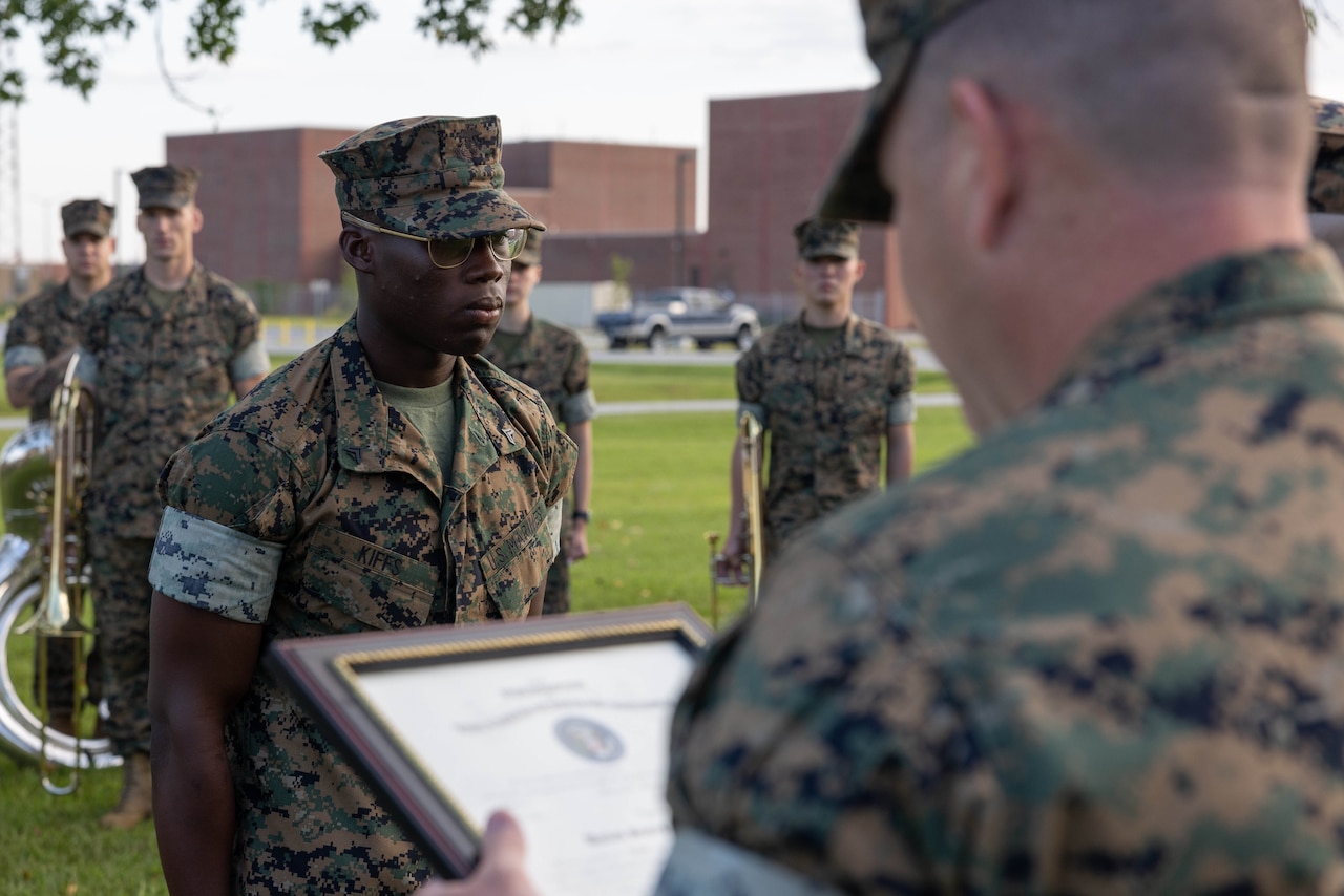 A Marine stands at attention as another holds a framed certificate while facing him on an outdoor field.