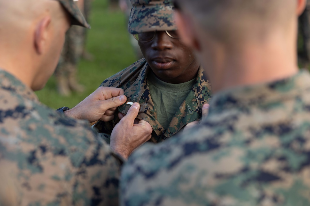 A Marine affixes a pin to the collar of another Marine's uniform as a third stands  by.