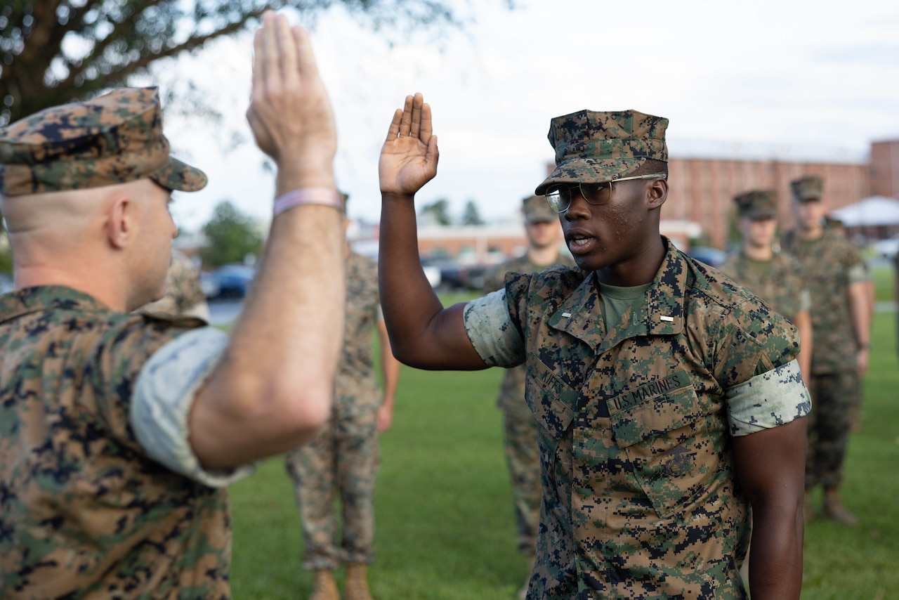 Two Marines face each other with right hands raised as others stand nearby in formation outdoors.