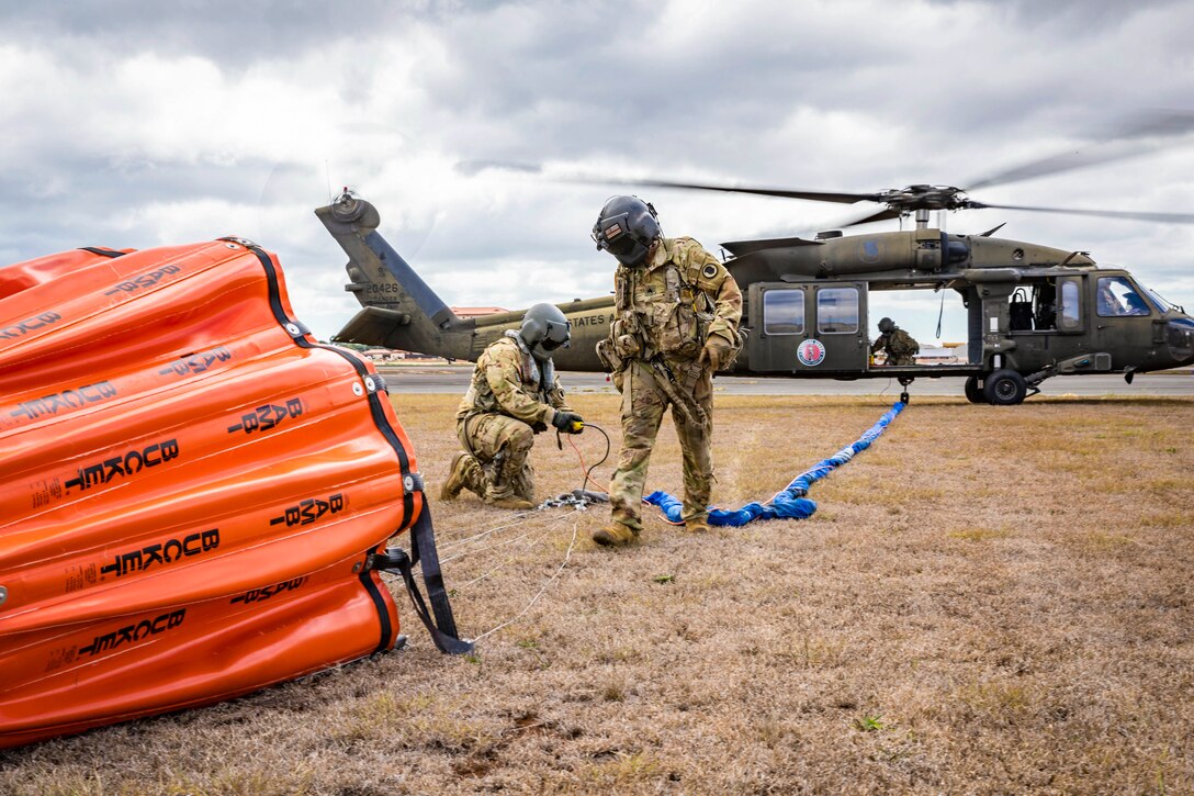 A soldier wearing a helmet walks toward an orange bucket as a fellow soldier kneels while connecting it to a helicopter parked in a field in the background.