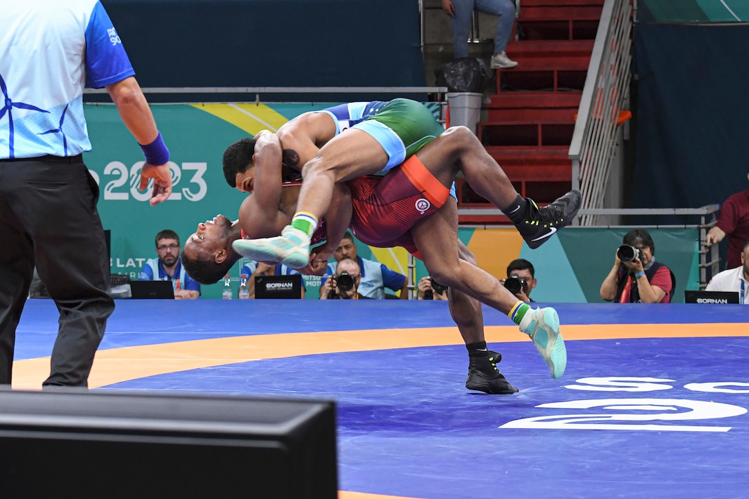 A soldier prepares to throw an opponent while falling backwards as judges watch and photographers capture the moment.