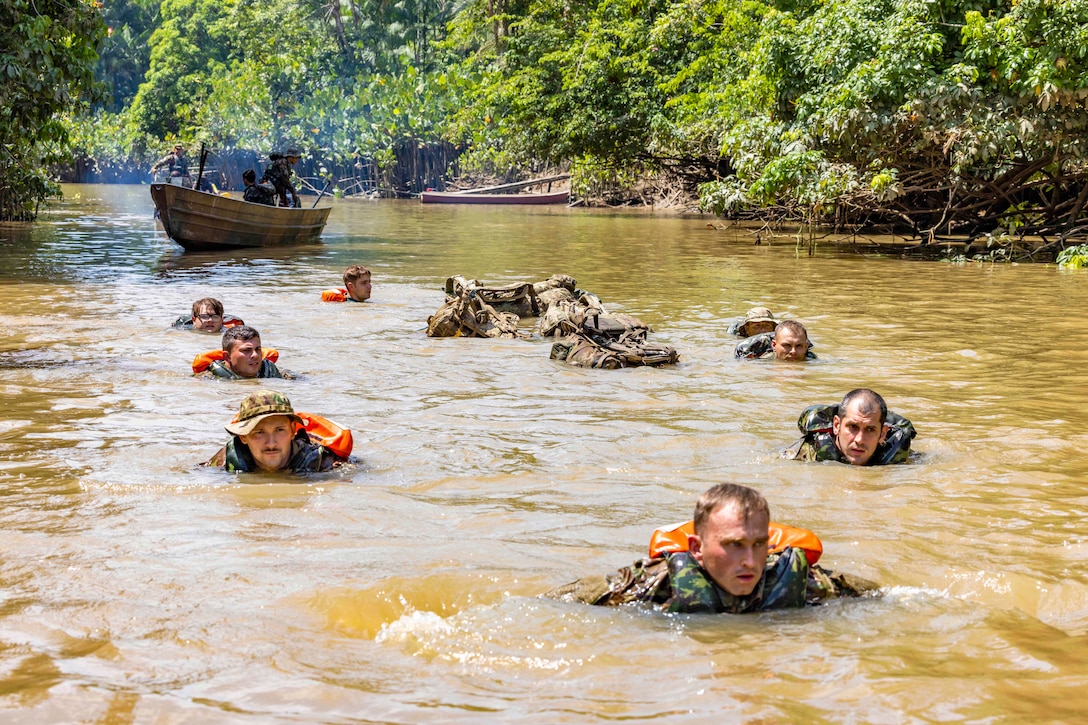 Soldiers wearing life vests move through a body of water as two small boats follow surrounded by greenery.