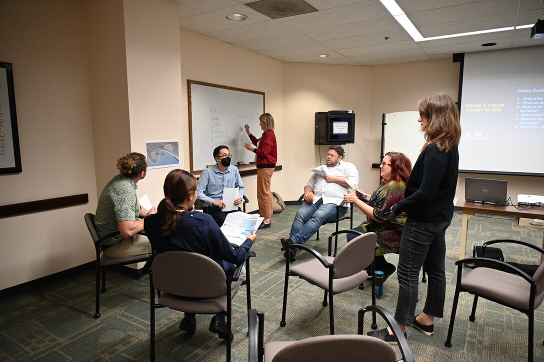 A white female stands to write on a white board as five white men and women sit in chairs as one white woman stands to their side and watches in a classroom setting.