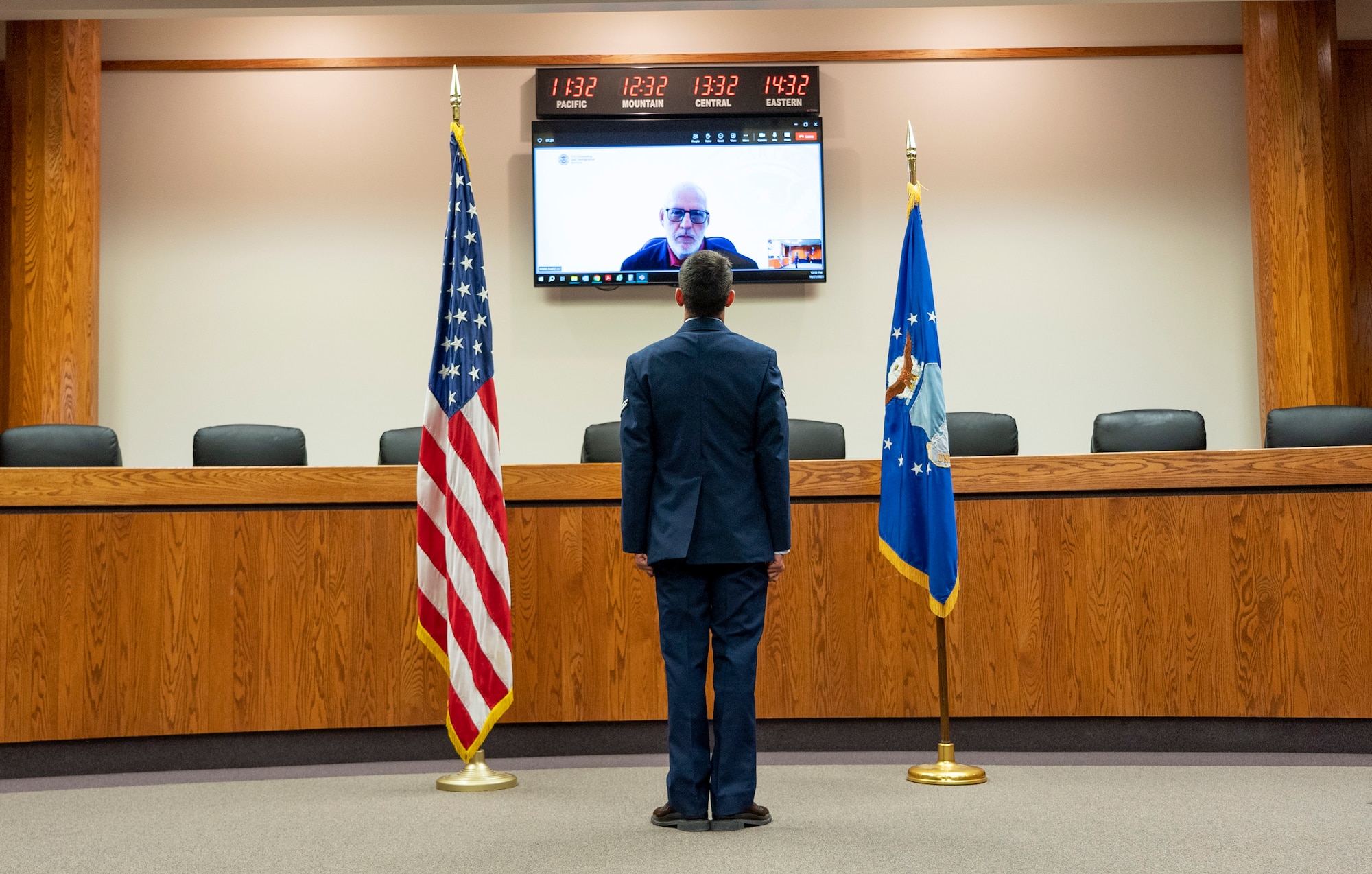 Airman 1st Class Minick standing at attention between the U.S. flag at left and U.S. Air Force flag at right looking up at a USCIS representative pictured on a television monitor.