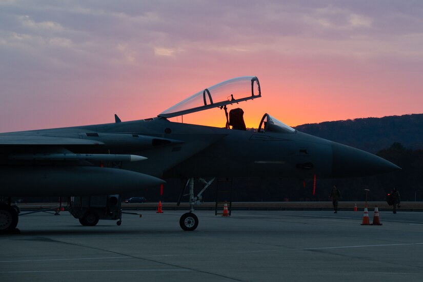 An aircraft with an open cockpit sits on the tarmac under a sunlit sky.