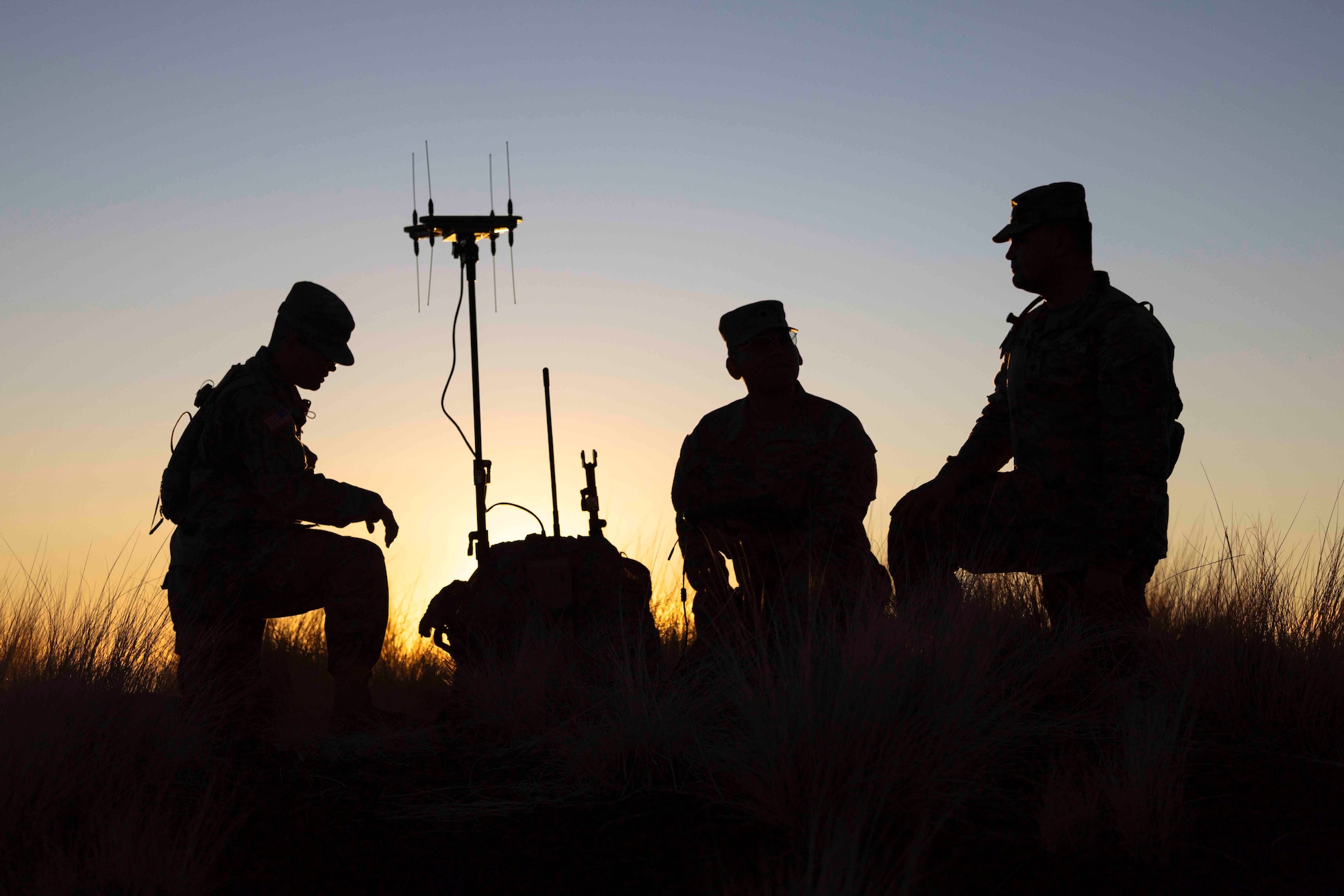 Three soldiers shown in silhouette kneel around a piece of equipment.