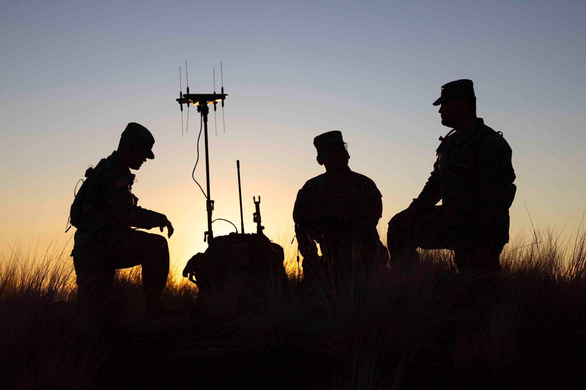 Three soldiers shown in silhouette kneel around a piece of equipment.