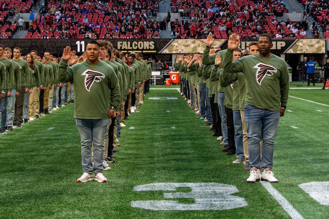 Rows of people stand on a football field while raising their right hand for an oath of enlistment ceremony.
