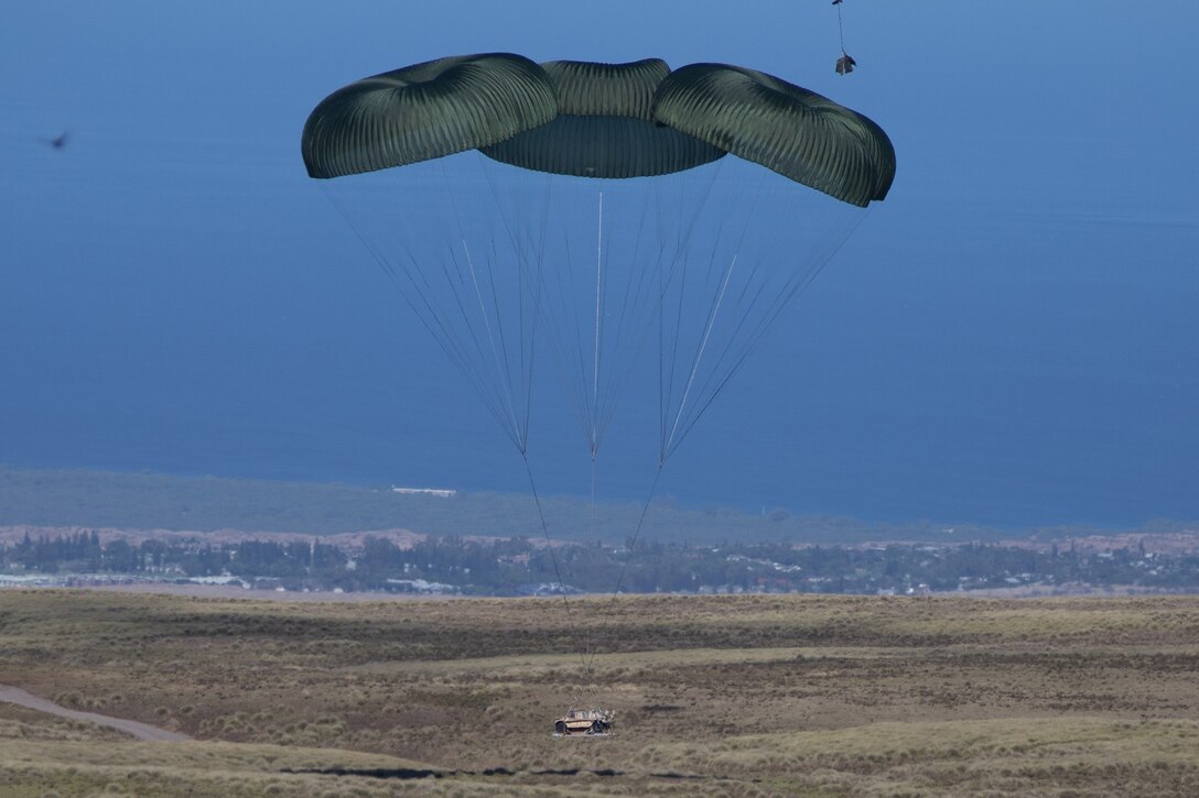 Service members conduct an airdrop of a military vehicle in an open field during daylight.