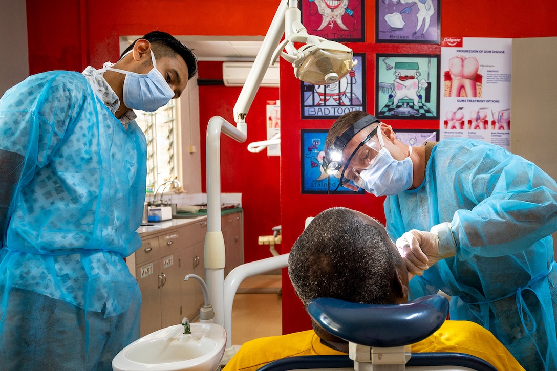 A dentist leans forward to examine a patient as another dentist watches.