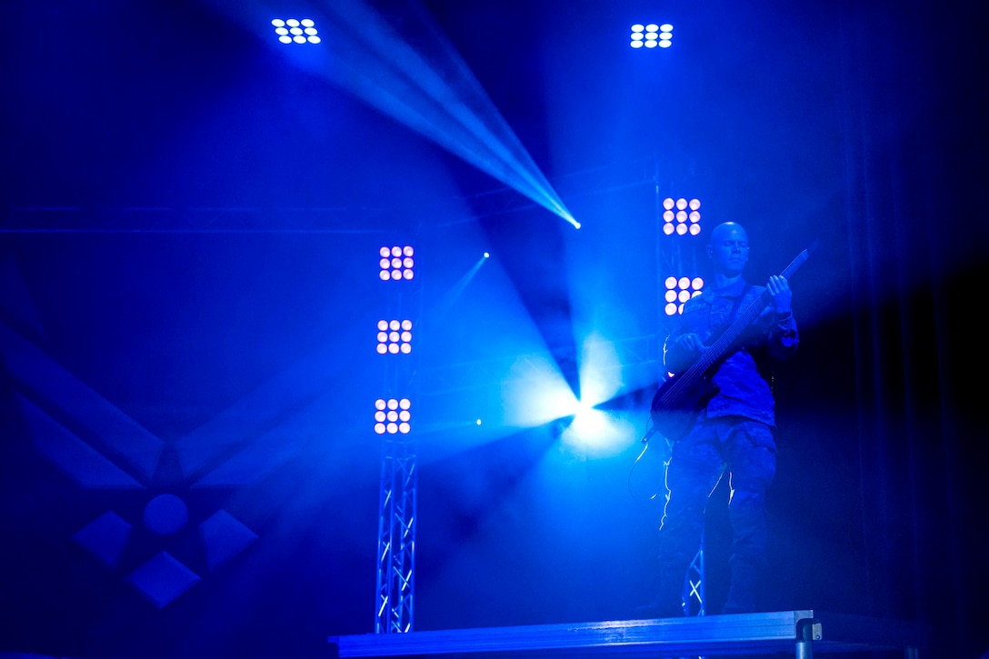 An airman illuminated by blue light plays a guitar on stage.