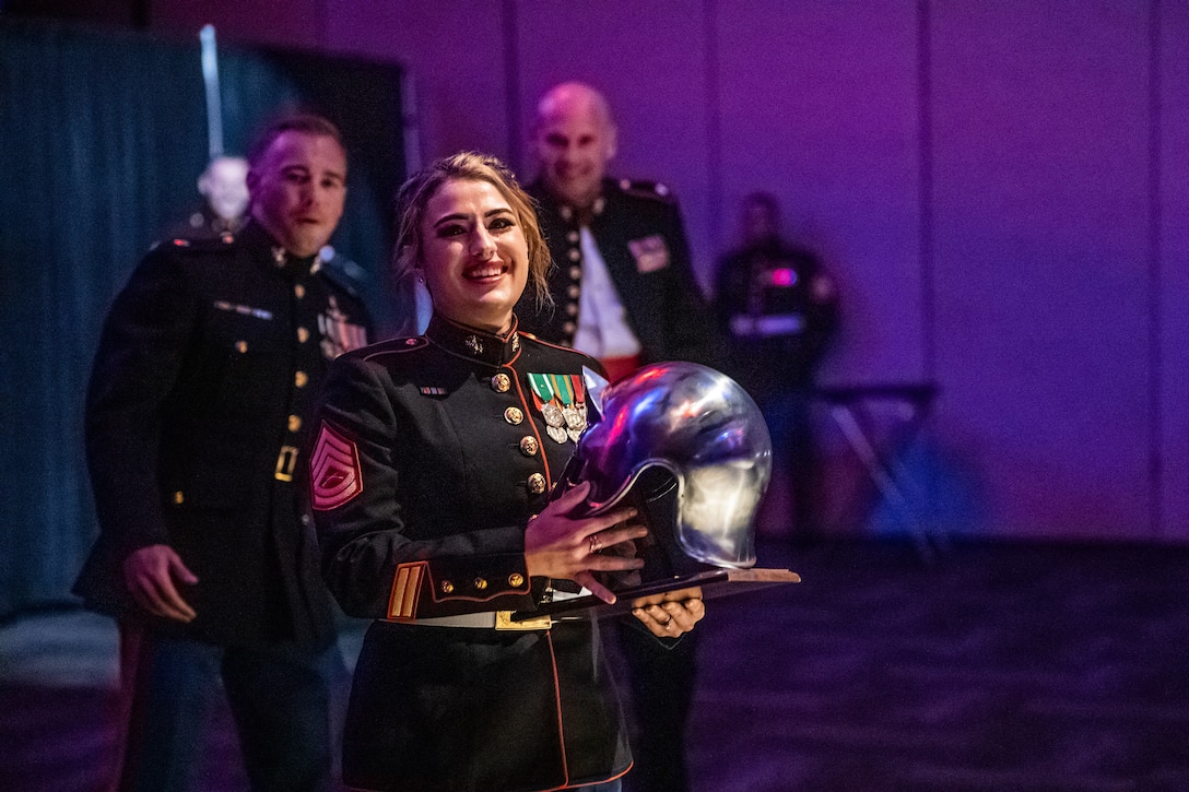 A Marine holds an award in a purple lit room as fellow Marines cheer in support in the background.