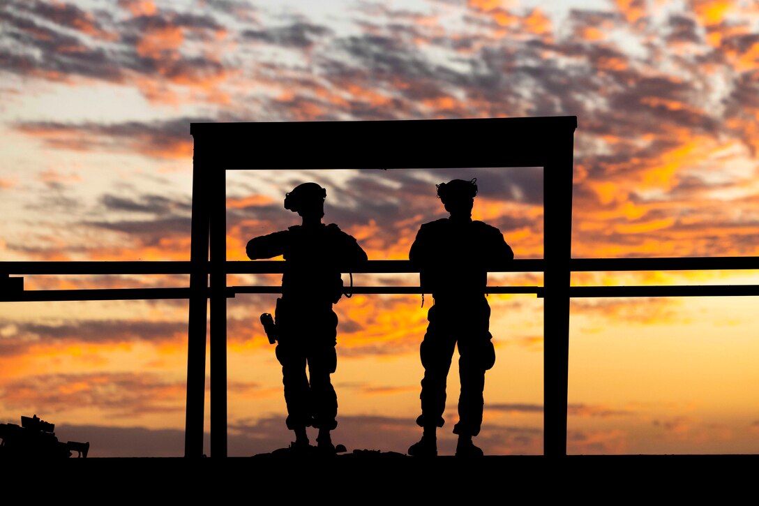 Marines stand in a structure under a sunlit sky as seen in silhouette.