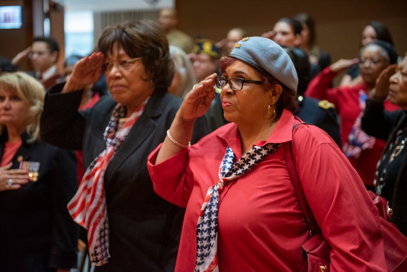 Two people in flag-patterned scarves tied as ties salute as others behind them do the same.