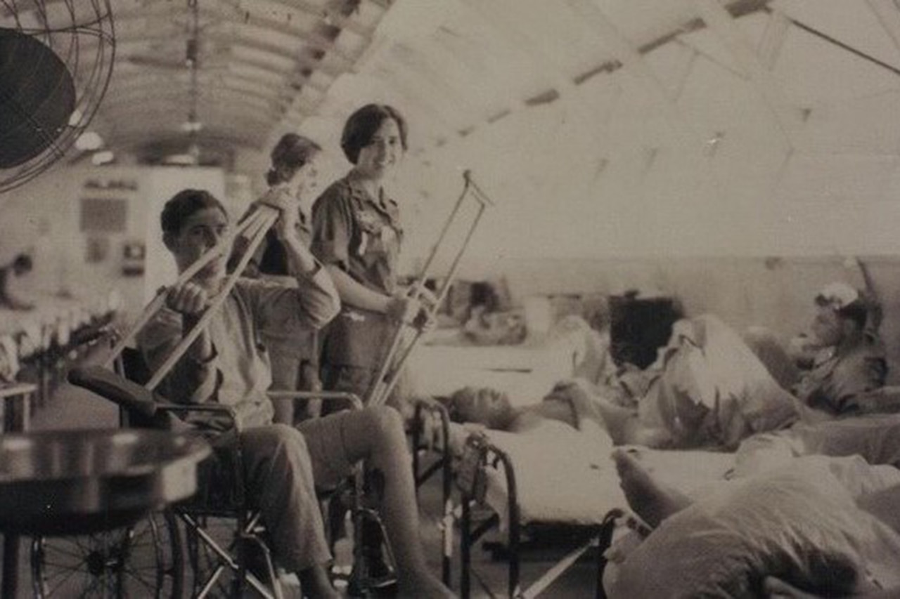 A person in a wheelchair in a room full of hospital beds holds up a crutch; another person beside them holds the other crutch.