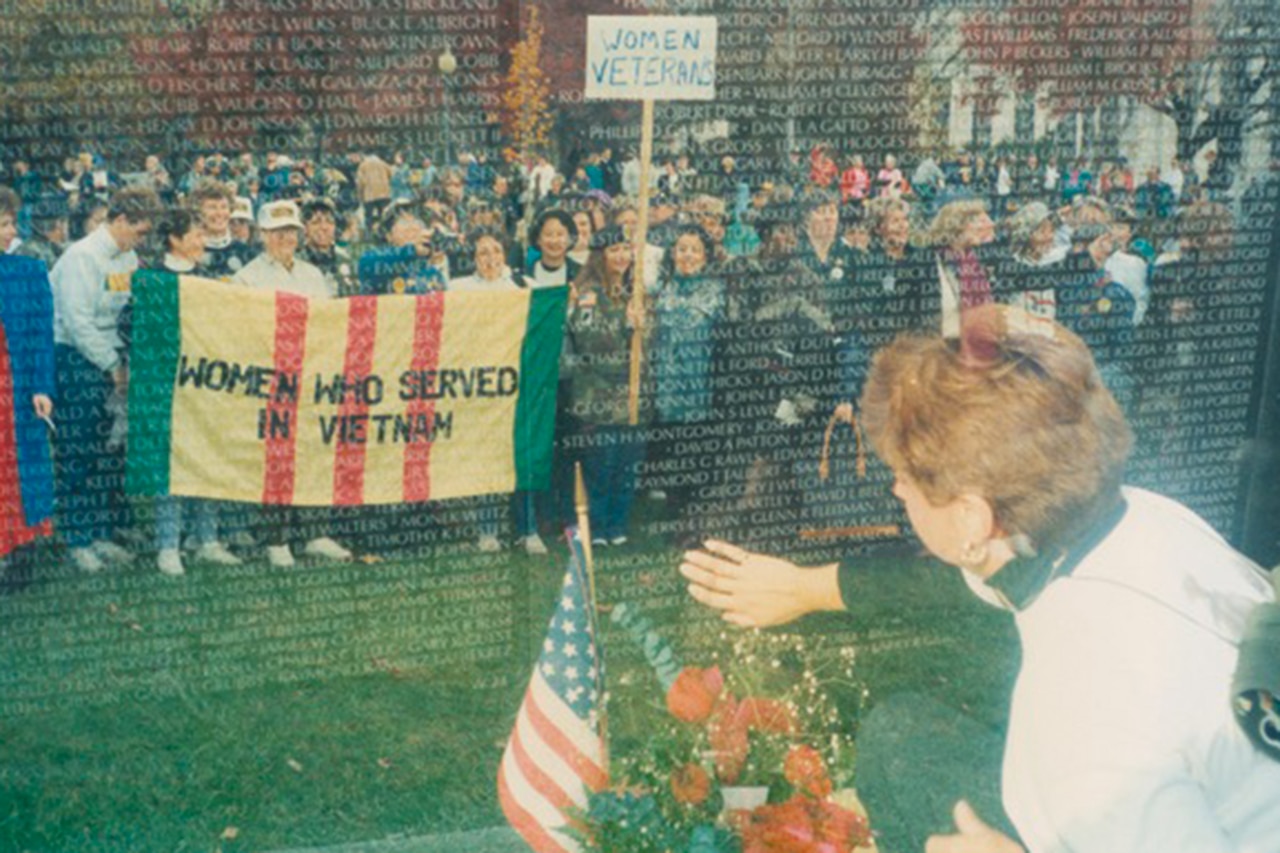 A person touches a name on a large wall of etchings. A reflection on the wall shows a large group of women standing behind them.