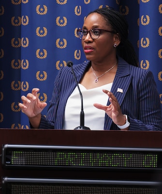 A Black woman stands at a podium speaking in front of a mic