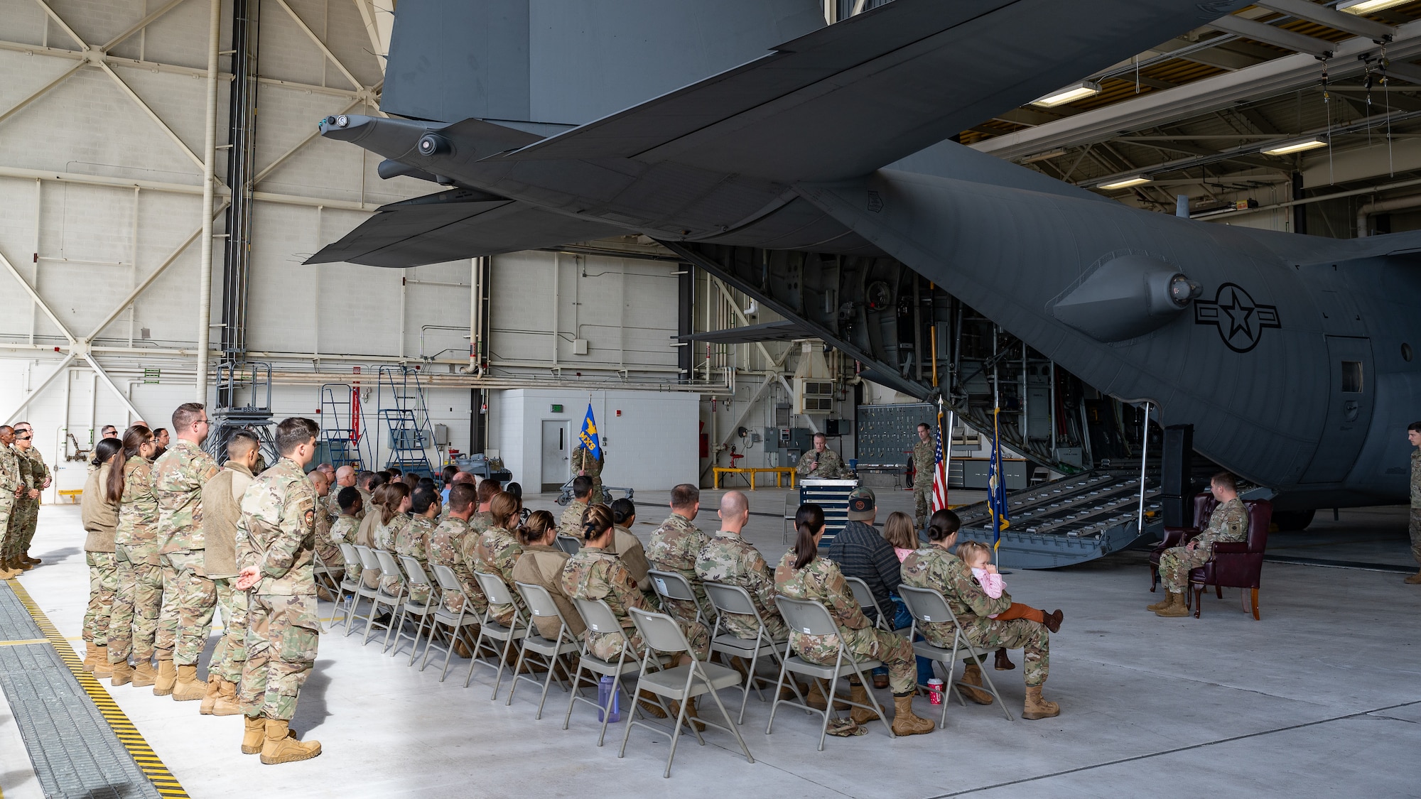A group of uniformed service members sit in chairs behind an open cargo bay of a C-130 aircraft as an audience for a ceremony.