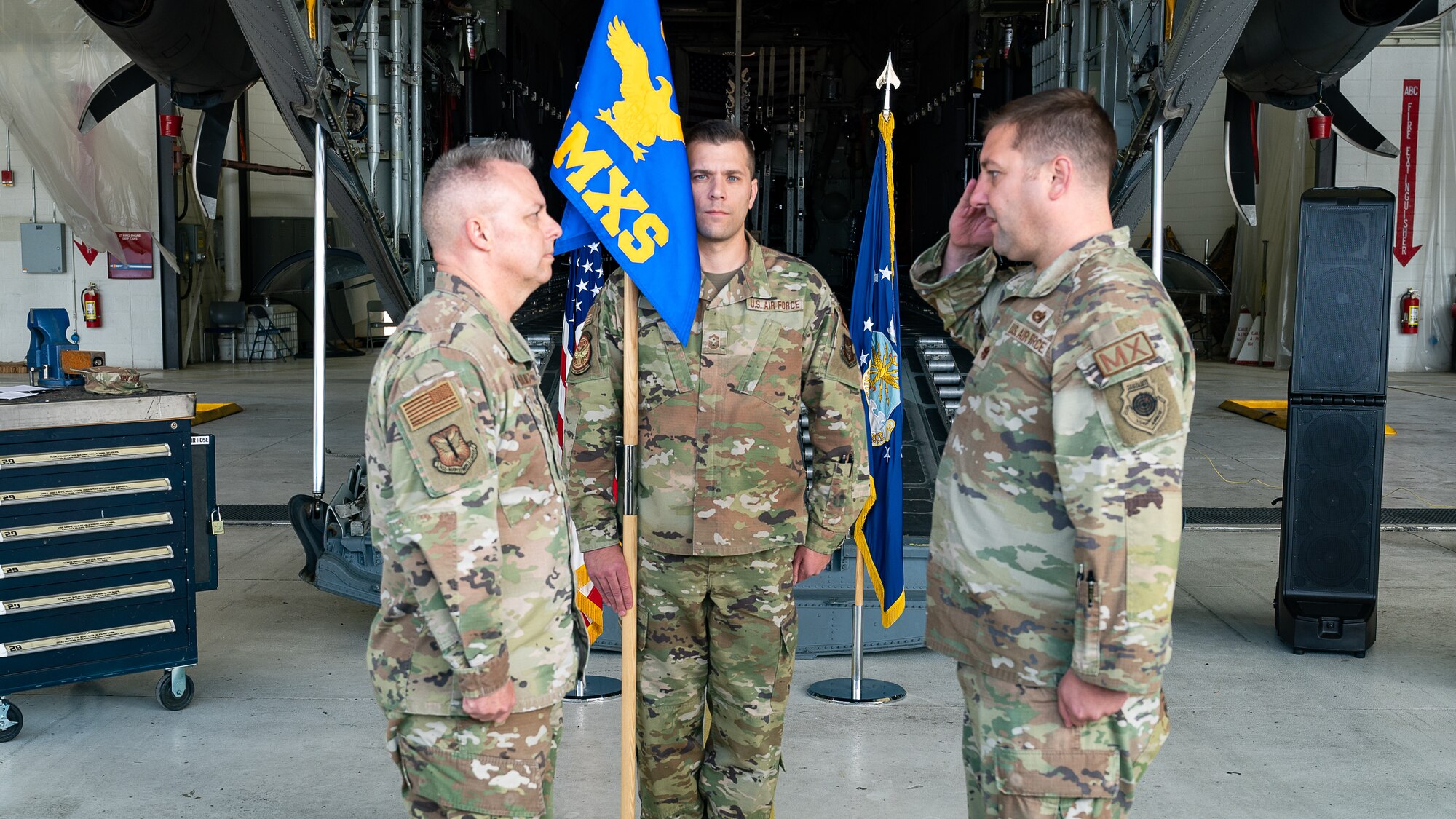 Two men in uniform salute each other while a third man in uniform holds a flag with "AMXS" embroidered on it, the group standing in front of a C-130 aircraft.