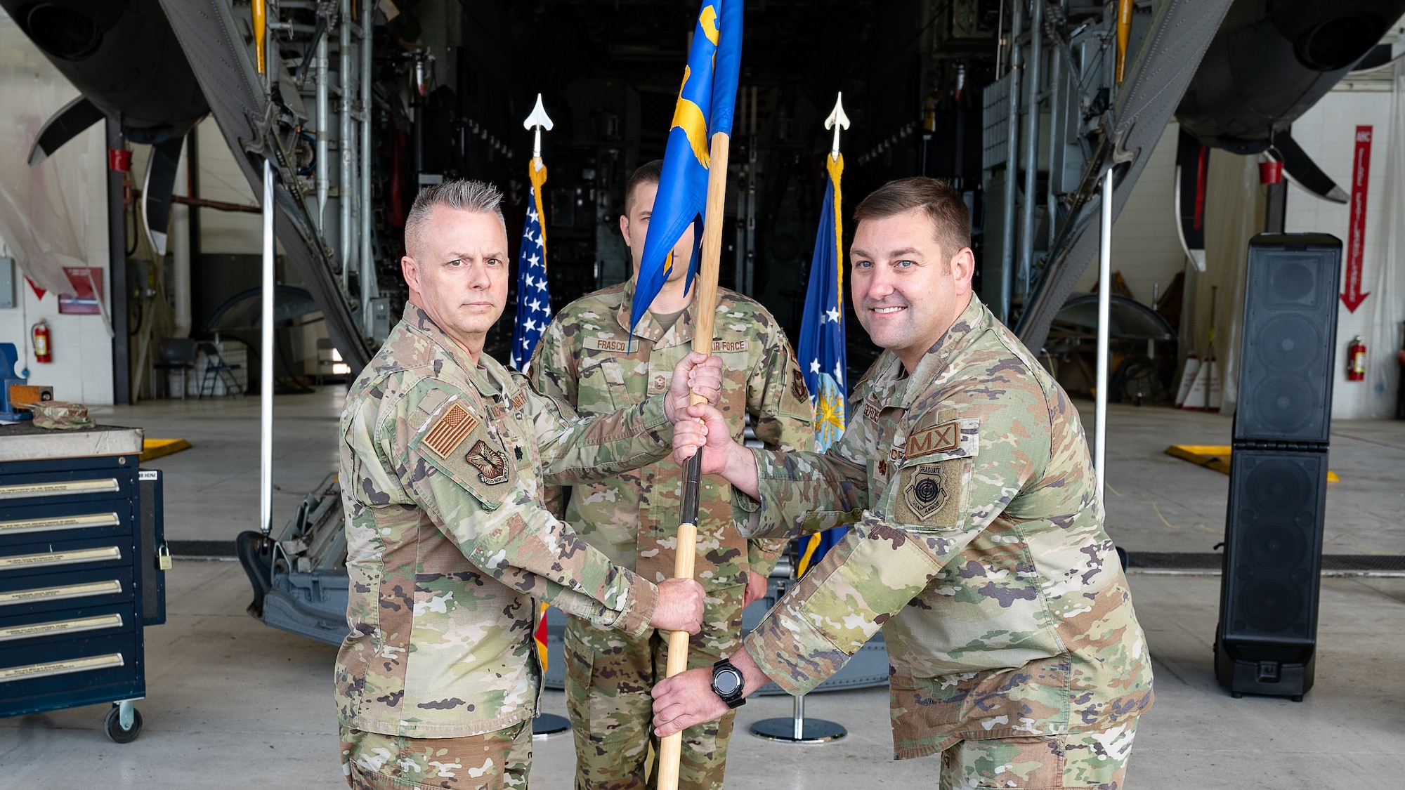 Two men in military uniform clasp a flagstaff affixed to a blue flag with a C-130 aircraft in the background.