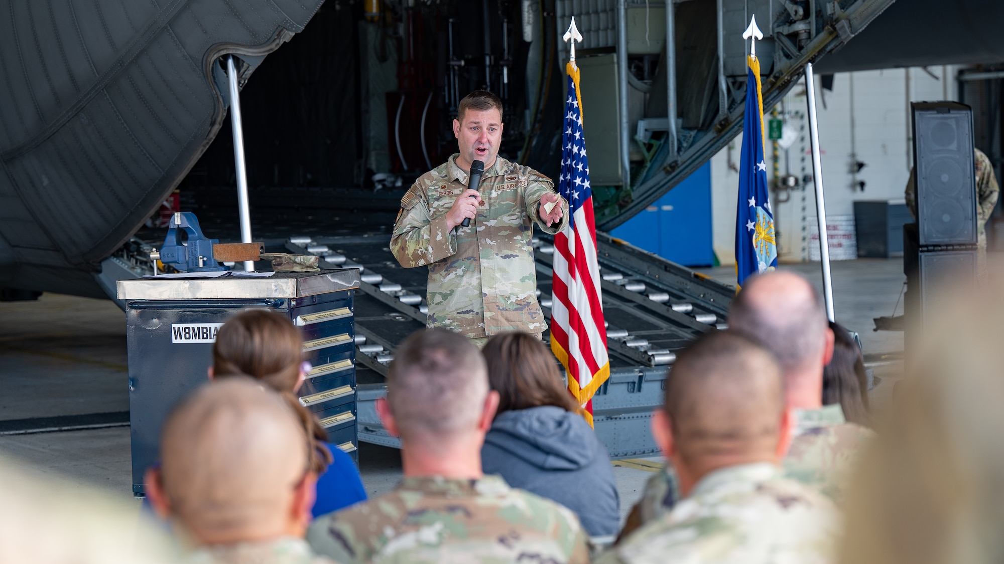 A man in military uniform next to a toolbox in front of an aircraft cargo bay speaks into a microphone at a crowd in the foreground.