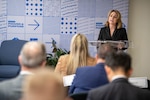 A woman in civilian attire stands at a lectern facing a seated audience.