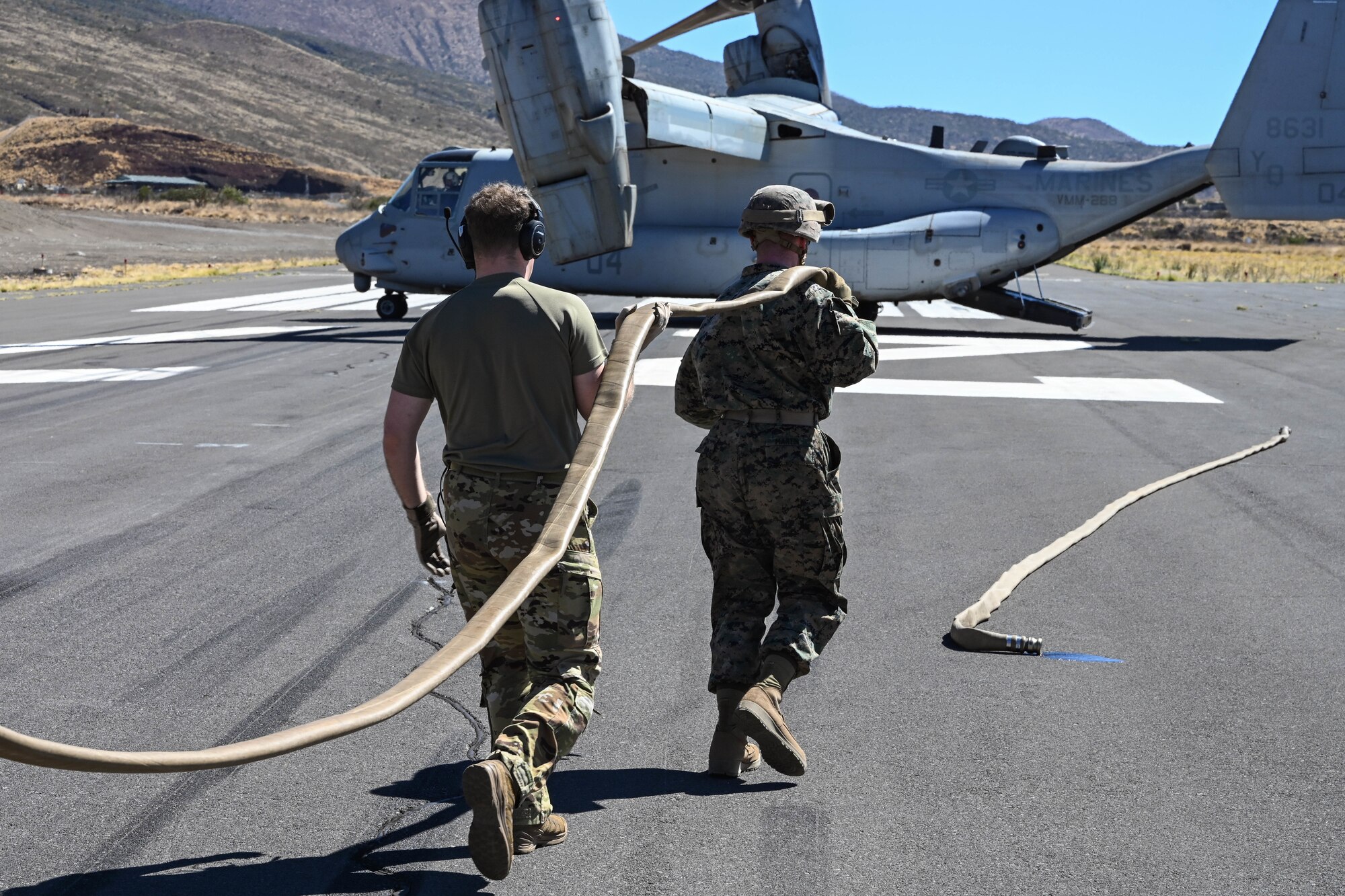 A U.S. Marine and a U.S. Airman carry a hose toward a MV-22B Osprey while conducting Forward Area Refueling Point operations.